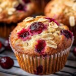 A golden-brown Homemade Cranberry Muffins in a white paper liner, topped with rolled oats and bursting cranberries, resting on a black cooling rack with more muffins and fresh cranberries in the background.
