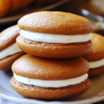 Homemade pumpkin whoopie pies with soft, golden-brown cakes and thick cream filling, stacked on a rustic plate with pumpkins in the background.