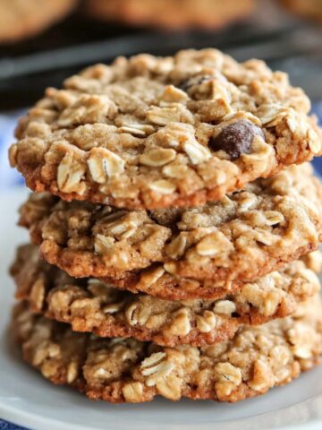 Stack of homemade Quaker-style quaker oatmeal cookie recipe on a white plate, packed with oats and raisins, with more cookies cooling in the background on a striped towel.