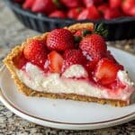 A slice of homemade strawberry cream cheese pie on a white plate with a gold rim, featuring a crumbly graham cracker crust, creamy white filling, and glossy fresh strawberries on top, set on a granite countertop with the full pie in the background.