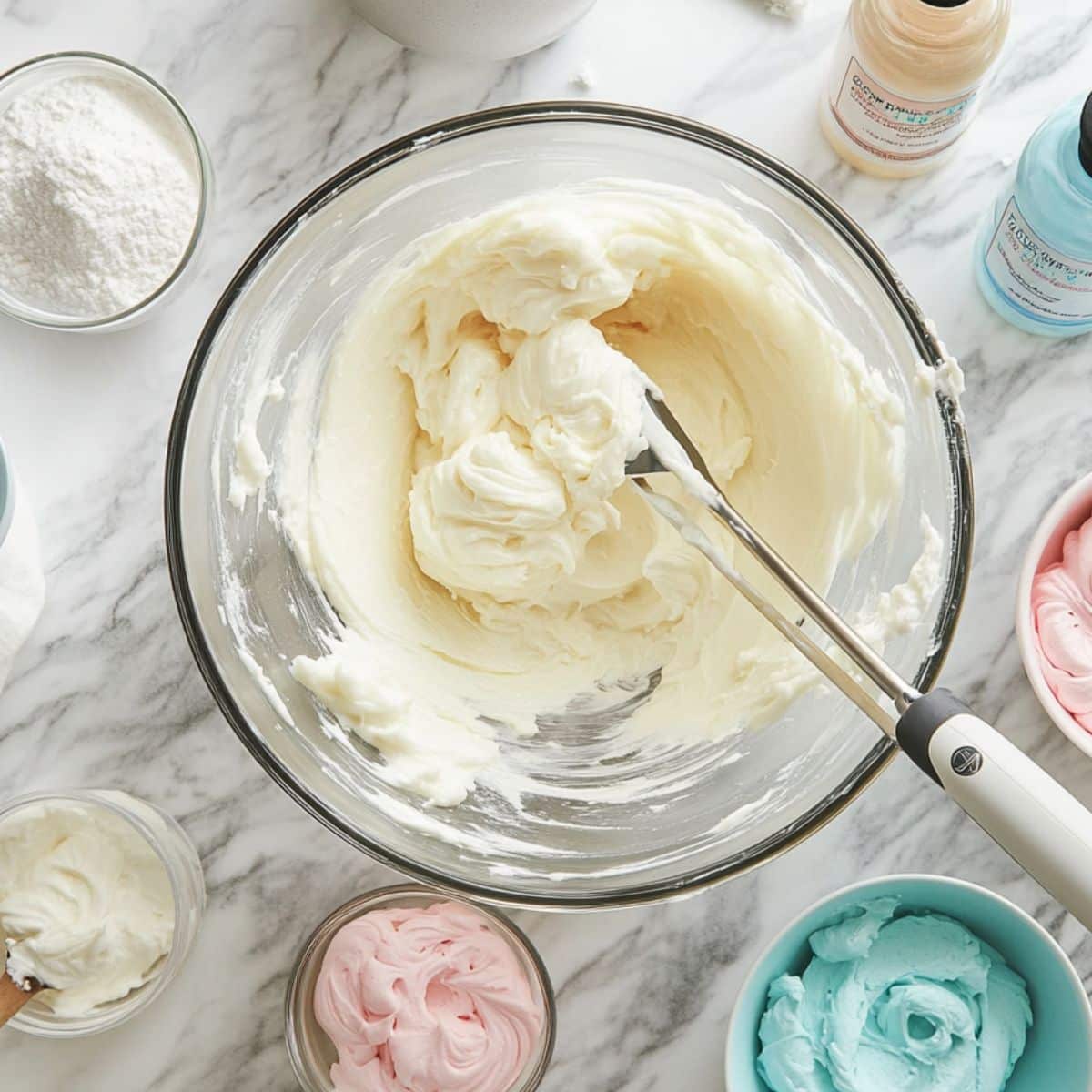 Homemade cotton candy frosting in white, pink, and blue, shown in bowls on a marble counter with food coloring and powdered sugar nearby.