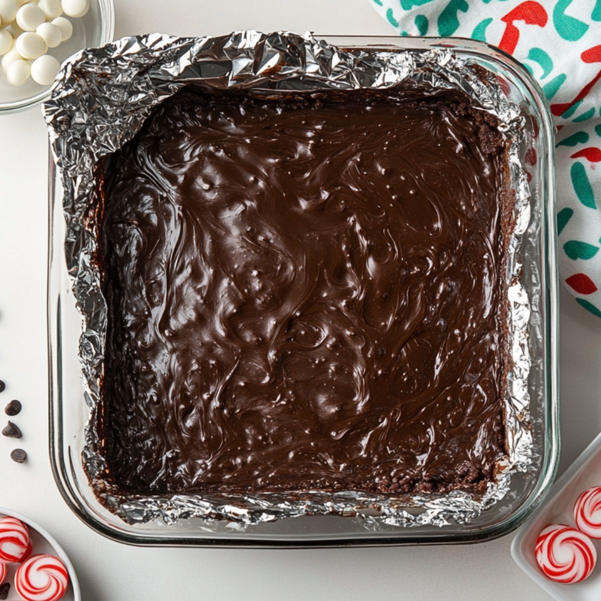 Glass baking dish with swirled chocolate brownie batter, lined with foil, surrounded by peppermint candies, chocolate chips, and a festive holiday towel.
