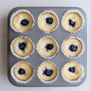 Unbaked lemon blueberry cupcake batter in a 9-cup metal muffin tray, each liner filled about two-thirds with a blueberry placed in the center.