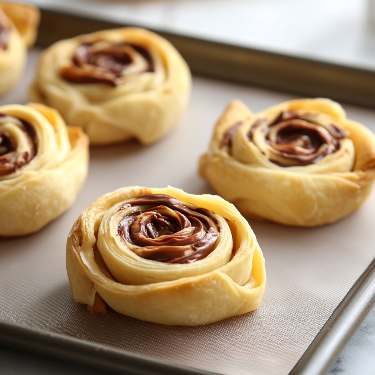 Golden, spiral-shaped Nutella puff pastries on a baking tray lined with a silicone mat, with flaky layers and glossy chocolate centers.