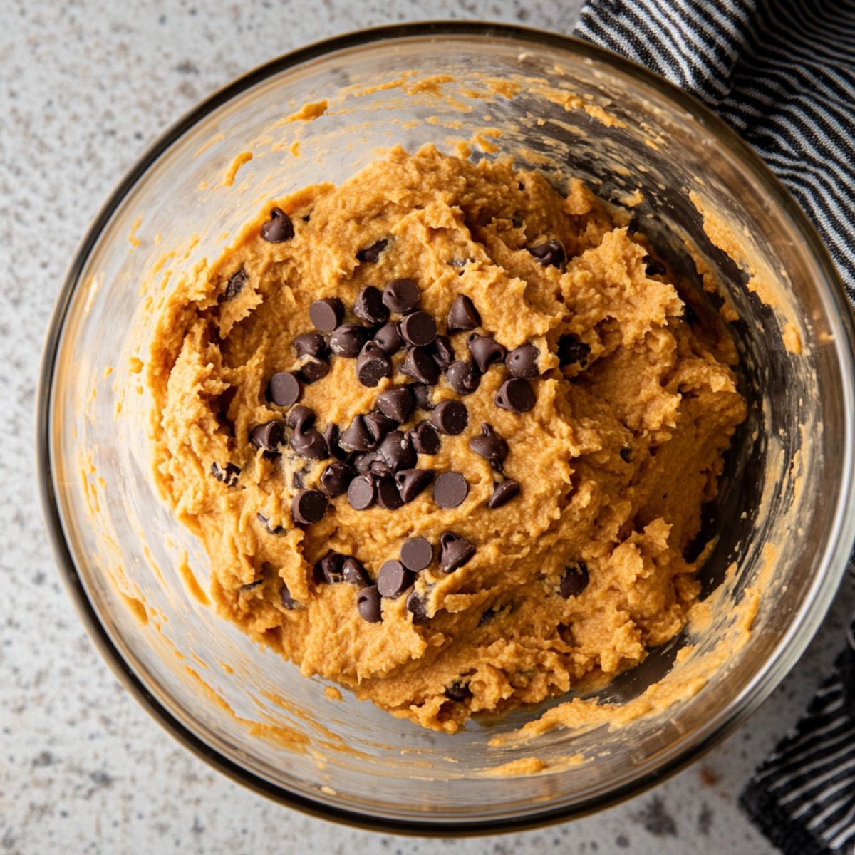 Pumpkin chocolate chip cookie dough in a glass bowl, with chocolate chips folded in, on a speckled countertop next to a striped towel.