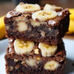 Homemade banana bread brownies stacked on a plate, with a fudgy chocolate base, baked-in banana chunks, and banana slices on top. Rich, moist, and freshly baked with a crackly top and ripe bananas in the background.