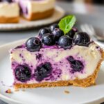Homemade blueberry cheesecake slice with a graham cracker crust, creamy filling swirled with blueberries, topped with fresh berries and a mint leaf on a white plate.