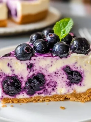 Homemade blueberry cheesecake slice with a graham cracker crust, creamy filling swirled with blueberries, topped with fresh berries and a mint leaf on a white plate.