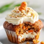 Homemade carrot cake cupcakes with cream cheese frosting, chopped nuts, and a fondant carrot on top, showing a moist interior and creamy filling after a bite.