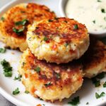Homemade golden-brown crab cakes with visible crab meat and herbs, served on a white plate with parsley garnish and a side of creamy dipping sauce.