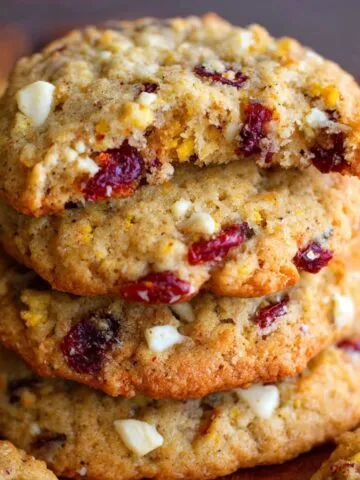 Stack of homemade cranberry orange cookies with white chocolate chunks, golden edges, and a soft, chewy center. Top cookie has a bite taken out, showing cranberries and zest inside.