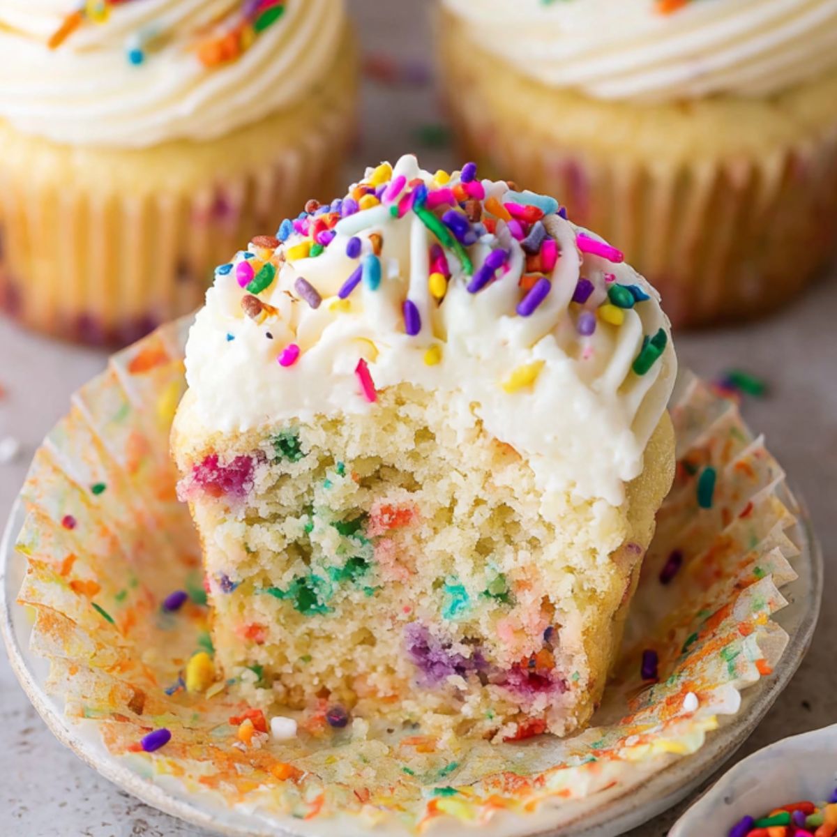 A bitten homemade funfetti cupcake with vanilla frosting and rainbow sprinkles, showing a colorful sprinkle-filled center. Surrounded by more cupcakes and scattered sprinkles on a kitchen counter.