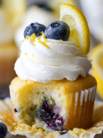Homemade Lemon Blueberry Cupcakes with cream cheese frosting, topped with fresh blueberries and lemon zest. One cupcake is bitten, showing juicy baked blueberries inside. Served on a white plate with scattered berries and a lemon in the background.