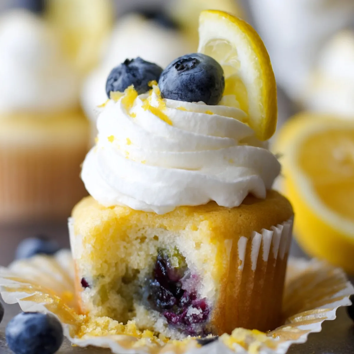 Homemade Lemon Blueberry Cupcakes with cream cheese frosting, topped with fresh blueberries and lemon zest. One cupcake is bitten, showing juicy baked blueberries inside. Served on a white plate with scattered berries and a lemon in the background.