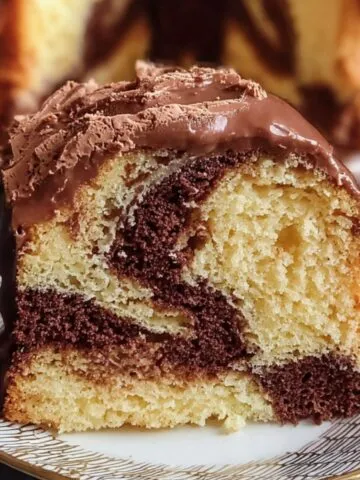 Homemade Marble Cake Recipe slice with rich chocolate and vanilla swirls, topped with glossy chocolate ganache and shavings, served on a decorative plate with the whole cake in the background.