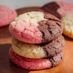Homemade Neapolitan cookies with pink, vanilla, and chocolate sections stacked on a wooden surface.