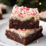 Homemade peppermint brownies topped with white frosting and crushed candy canes, stacked on a white plate with a festive, blurred Christmas background.