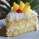 A slice of homemade pineapple coconut cake with two layers of moist yellow sponge, creamy frosting, and shredded coconut on top. Garnished with fresh pineapple wedges and a cherry, served on a ceramic plate with a soft, natural background.