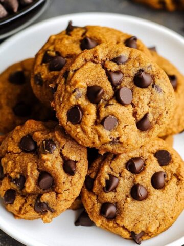 Homemade pumpkin chocolate chip cookies stacked on a white plate, golden and soft with melty chocolate chips and a rustic, cozy look.