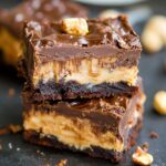 Stack of homemade Snickers brownies with fudgy chocolate base, creamy nougat center, and glossy ganache topping, garnished with peanuts and caramel, on a dark surface with scattered crumbs.