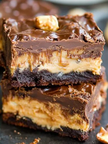 Stack of homemade Snickers brownies with fudgy chocolate base, creamy nougat center, and glossy ganache topping, garnished with peanuts and caramel, on a dark surface with scattered crumbs.