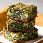 Stack of homemade spinach brownies with golden edges and a moist, green spinach-filled center on a white plate, set against a warm kitchen background.