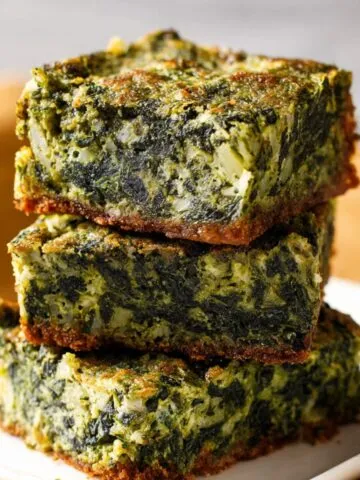 Stack of homemade spinach brownies with golden edges and a moist, green spinach-filled center on a white plate, set against a warm kitchen background.