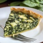 A slice of homemade spinach pie recipe with a golden flaky crust and creamy spinach-feta filling on a white plate, with a fork and fresh spinach leaves in the background.