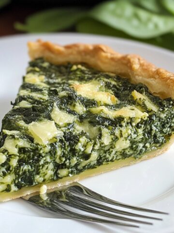 A slice of homemade spinach pie recipe with a golden flaky crust and creamy spinach-feta filling on a white plate, with a fork and fresh spinach leaves in the background.