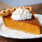 Homemade slice of sweet potato pie with flaky crust, whipped cream, and cinnamon on a white plate.