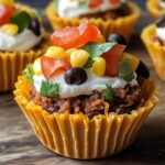 Homemade Taco Cupcakes in a yellow liner, topped with ground beef, sour cream, tomatoes, corn, black olives, and cilantro on a wooden surface.