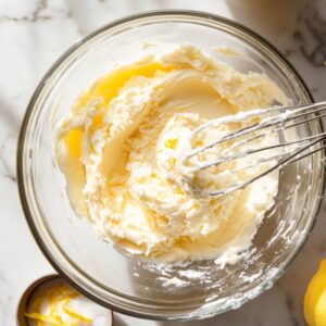 Whipped cream cheese lemon frosting in a glass bowl with a metal whisk, set on a marble surface with lemon zest and a fresh lemon nearby.