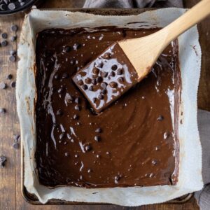 A parchment-lined baking pan filled with glossy banana chocolate brownie batter, being spread evenly with a wooden spatula, with chocolate chips visible on the surface.