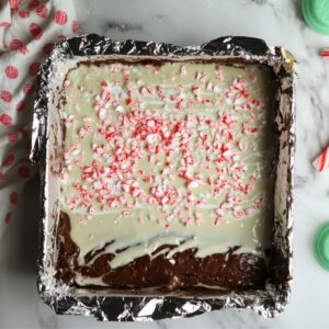 Pan of peppermint brownies topped with melted white chocolate and crushed candy canes, lined with foil and set on a marble counter with festive baking items around.