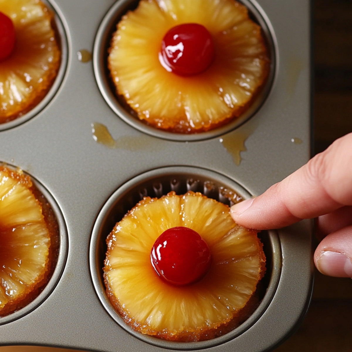 A hand gently presses a caramelized pineapple ring and cherry topping into a freshly baked cupcake still inside a muffin tin, surrounded by other golden brown pineapple upside-down cupcakes.