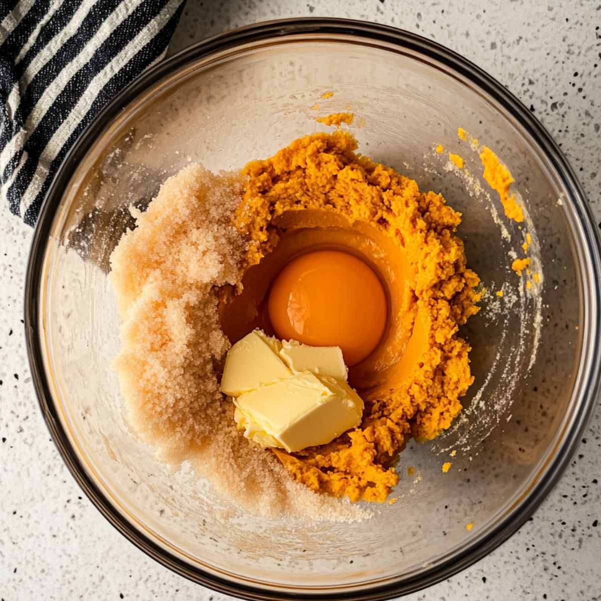 Unmixed pumpkin cookie ingredients in a glass bowl—pumpkin puree, a raw egg, brown sugar, and chunks of butter—on a speckled counter next to a striped kitchen towel.