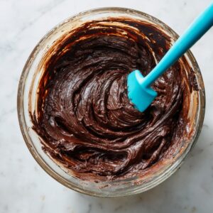 Thick chocolate brownie cookie dough in a glass bowl with a blue silicone spatula on a marble countertop. The dough is dark, glossy, and rich, swirled with visible texture from mixing.