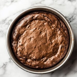 Freshly baked flourless chocolate cake in a round metal pan with a cracked, set top and slightly domed center, cooling on a marble surface.