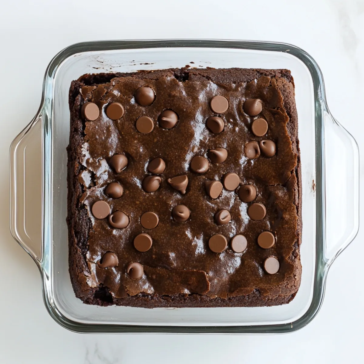 Freshly baked black bean brownies in a square glass dish, topped with melty chocolate chips on a glossy, crackled surface.