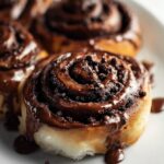 Homemade chocolate cinnamon roll, golden and gooey with melted chocolate dripping down the sides, topped with chocolate shavings on parchment over rustic wood.