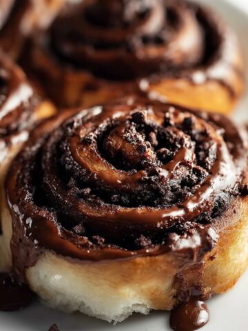 Homemade chocolate cinnamon roll, golden and gooey with melted chocolate dripping down the sides, topped with chocolate shavings on parchment over rustic wood.