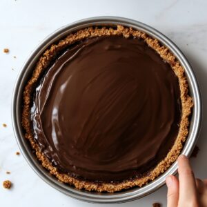 Homemade s’mores pie with a smooth, glossy chocolate ganache layer in a graham cracker crust, set in a metal pie pan on a marble surface, with a hand gently holding the edge.