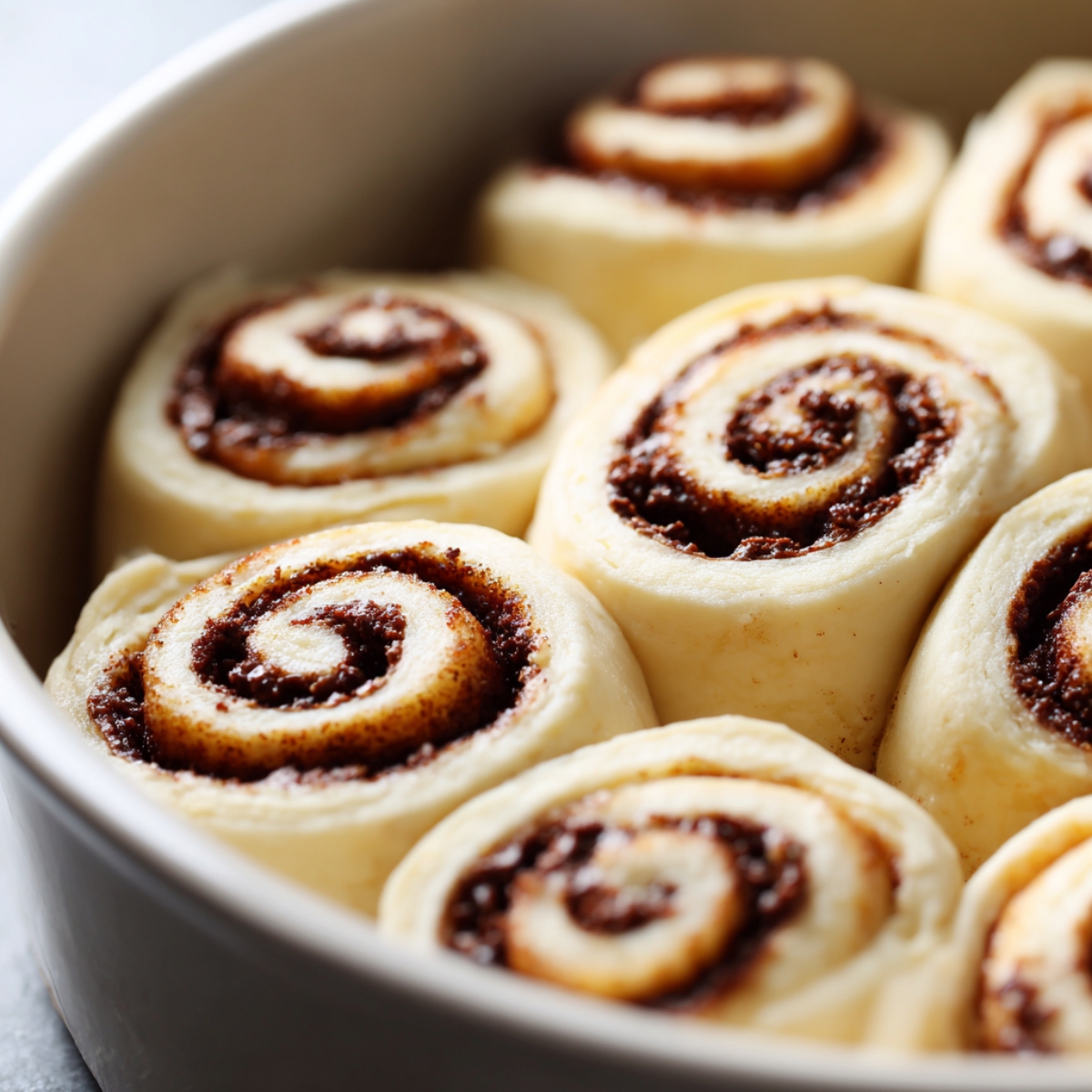 Close-up of unbaked chocolate cinnamon rolls arranged in a round baking dish, showing smooth dough spirals with rich cocoa filling, ready for proofing or baking.