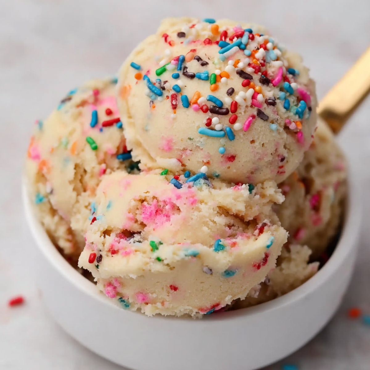 Scoops of edible sugar cookie dough with rainbow sprinkles in a white bowl, with a gold spoon on a light background.