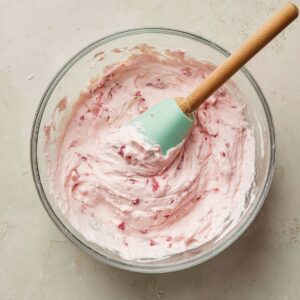 Homemade strawberry cheesecake filling in a glass mixing bowl with a mint green spatula, showing a fluffy pink texture with visible strawberry pieces.