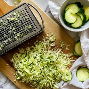 Grated zucchini on a wooden cutting board with a box grater, zucchini slices, and a kitchen towel nearby—prepping for chocolate zucchini cake.