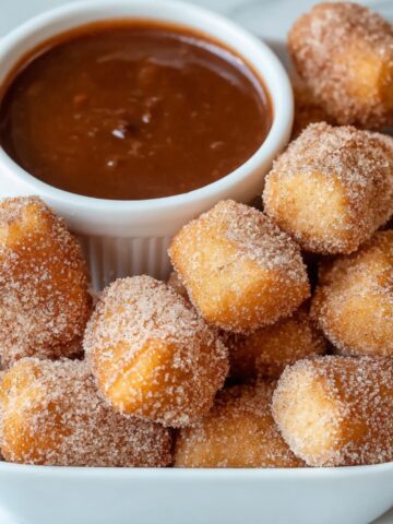 Homemade air fryer churro bites coated in cinnamon sugar, served in a white square dish with a ramekin of thick chocolate dipping sauce.