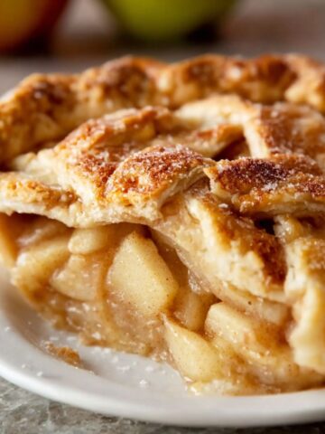Close-up of a slice of homemade apple pie recipe with a golden, flaky lattice crust, filled with tender, cinnamon-spiced apple slices, lightly dusted with sugar.