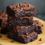 A stack of rich, homemade black bean brownies on a rustic ceramic plate, with a moist, fudgy texture, cracked tops, and melted chocolate chips scattered on and around them.