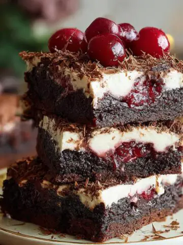 Homemade Black Forest brownies stacked on a plate, with layers of fudgy chocolate, cherry filling, whipped cream, and topped with cherries and chocolate shavings.